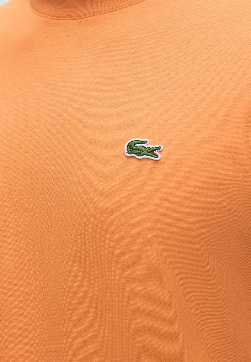 Oranje stof met een klein groen geborduurd krokodillenlogo met rode details op het bovenste borstgedeelte.