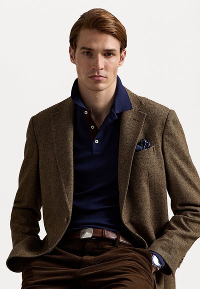 Brown herringbone blazer over a navy polo shirt, with a pocket square. Brown corduroy pants and a silver-buckled brown belt complete the look.