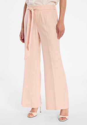 Frau trägt hoch taillierte, weit geschnittene hellrosa Hose mit passendem Stoffgürtel und weißen, offenen High-Heel-Schuhen.