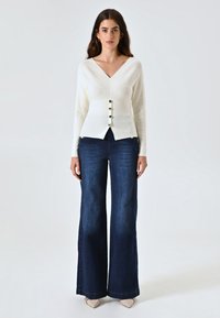 Maglione bianco a collo a V con dettagli di bottoni, indossato con jeans a zampa d'elefante blu scuro. Tessuto morbido e forma aderente, abbinato a scarpe di colore chiaro.