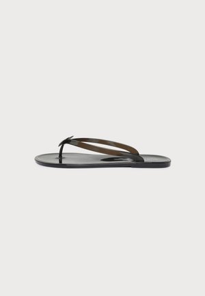 Sandale plate noire de type tong, avec des brides brillantes et un petit détail en forme de nœud, présentée de profil sur un fond clair uni.