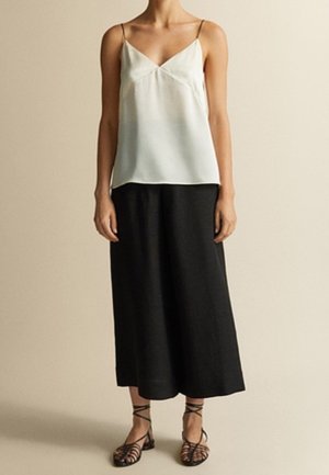 Donna che indossa una canotta bianca senza maniche con scollo a V, pantaloni neri a gamba larga e corti e sandali neri piatti con cinturini su uno sfondo neutro.