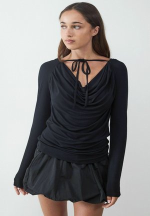 Schwarzes Langarmoberteil mit drapiertem Ausschnitt und Bindeband am Rücken; kombiniert mit einem schwarzen, ausgestellten Rock, beide aus glattem Stoff.