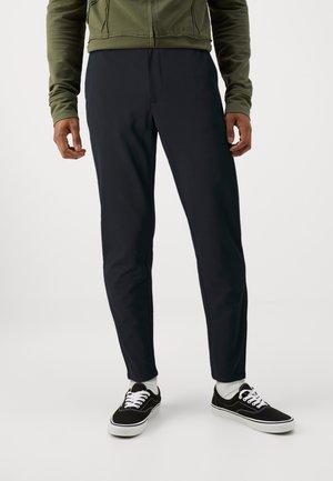 Man draagt zwarte strakzittende broek, zwart-witte sneakers, witte sokken en een olijfgroene jas, staand tegen een witte achtergrond.