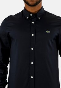 Camicia button-up blu navy in cotone, con bottoni bianchi e un piccolo logo di coccodrillo ricamato in verde sul lato sinistro del petto.