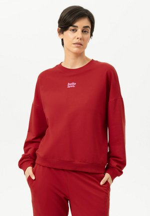 Femme portant un sweat-shirt rouge et un pantalon, texte sur le sweat-shirt : « hello love », mains dans les poches, expression neutre, fond blanc uni.