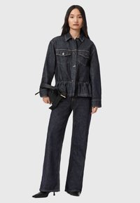 Donna che indossa una giacca in denim scuro e jeans a gamba larga, con in mano una borsa a cintura nera, in piedi davanti a uno sfondo chiaro e semplice.