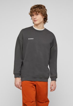 Giovane uomo con capelli ricci che indossa una felpa Mammut grigio scuro e pantaloni arancioni vivaci, in piedi su uno sfondo chiaro e semplice.