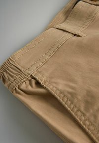 Beez värvi kangast lühikesed püksid elastse vöökoha, detailse õmbluse ja lamedate esikottidega. Tekstuur näeb välja pehme ja sile.