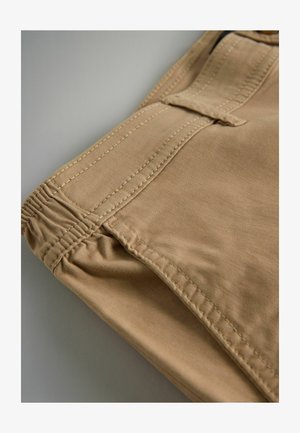 Smėliniai audinio šortai su elastinga juostele, išsamiomis siūlėmis ir plokščiu priekiu kišenėje. Audinys atrodo minkštas ir lygus.