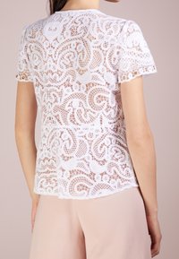 Biała haftowana koronkowa bluzka z krótkim rękawem z misternymi kwiatowymi wzorami, noszona z jasnoróżowymi spodniami, pokazana od tyłu na jasnym tle.
