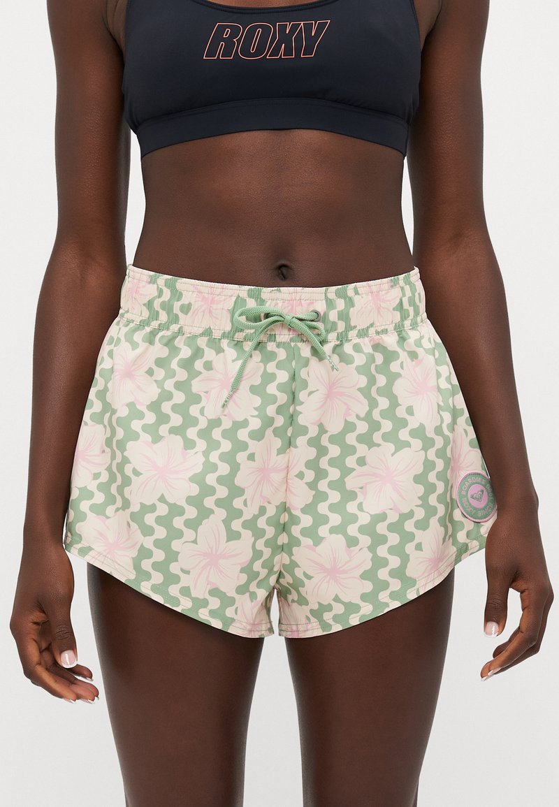Swim shorts featuring a pastel floral pattern on a green wavy background, with an elastic waistband and drawstring. Side patch detail included.