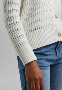 Krämfärgad stickad cardigan med ett mönstrat tyg, ribbad fåll och tre rundade knappar; matchad med blå denimjeans.