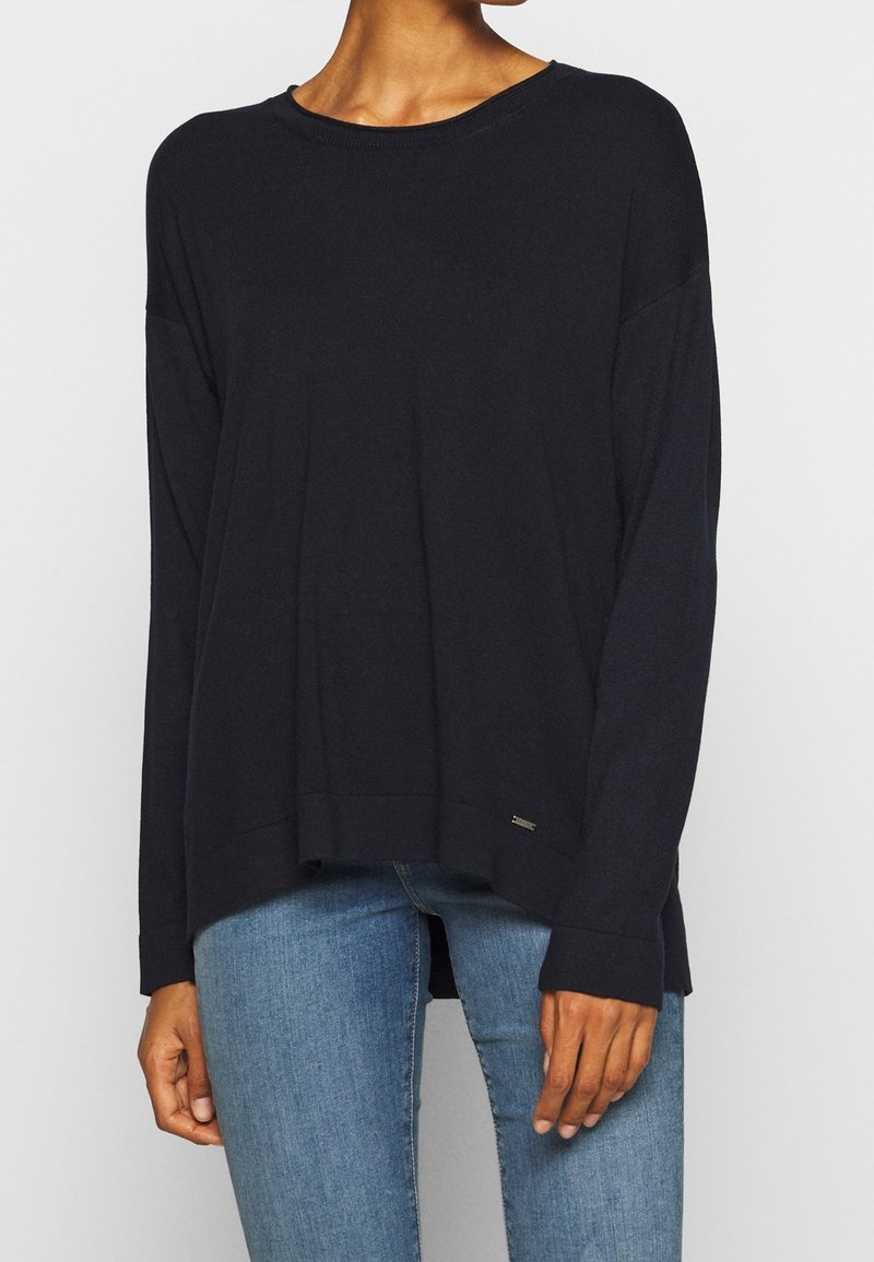 Femme portant un pull noir ample à manches longues avec un col rond et un jean en denim bleu, debout devant un fond uni.