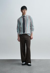 Pruhovaná košile na knoflíky v světle modré, červené a bílé barvě, doplněná hnědými širokými kalhotami a černými botami s bílými detaily.