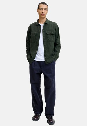 Man staat en draagt een donkergroene overhemdenjas met knopen, een wit T-shirt, losse marineblauwe broek en donkerbruine leren schoenen tegen een effen achtergrond.