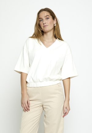 Mujer con cabello largo vistiendo una camiseta blanca de manga corta con escote en V y pantalones beige, de pie contra un fondo blanco liso.