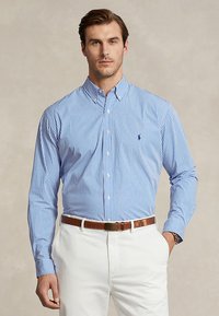 Férfi kék-fehér csíkos, galléros gombos ingben, fehér nadrágban, barna övvel és órával, semleges háttér előtt áll.