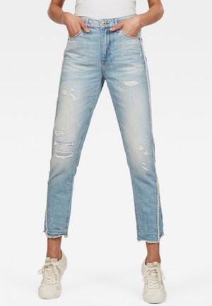 Donna che indossa jeans azzurri strappati con orlo grezzo e sneakers bianche, in piedi con le mani nelle tasche.