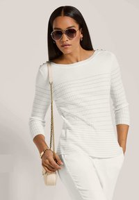 Weißer, gerippter Pullover mit langen Ärmeln, strukturiertem Design und Knopfdetails an den Schultern. Kombiniert mit weißen Hosen und einer hellen Handtasche.