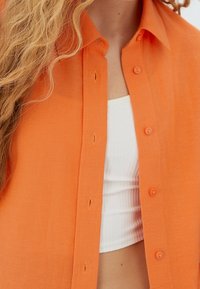 Vrouw met lang blond haar, gekleed in een open oranje shirt over een wit geribbeld crop top, torso zichtbaar.
