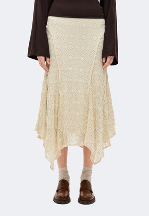 Jeune personne aux cheveux courts et foncés porte un haut à manches longues marron, une jupe midi crème texturée avec un ourlet en pointe, des chaussettes beiges et des mocassins marron.