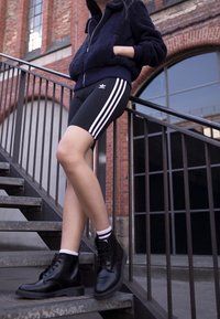 Black bicycle shorts with three white stripes, paired with a navy fleece zip-up jacket and black lace-up boots, on a metal staircase.