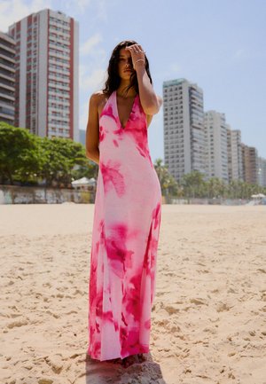 Žena u ružičastoj i crvenoj halter haljini stoji bosonoga na sunčanoj plaži s visokim zgradama i drvećem u pozadini.