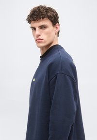 Νεαρός άνδρας με καστανά σγουρά μαλλιά που φοράει μπλε σκούρο φούτερ, κοιτώντας πάνω από τον ώμο του, με φόντο ένα απλό ανοιχτόχρωμο φόντο.