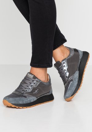 Voeten met grijs-zwarte sneakers met gumzolen, gecombineerd met zwarte nauwsluitende broek op een lichtgrijze vloer.