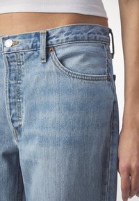 Jeans de mezclilla azul claro con una cintura alta, diseño de cinco bolsillos y herrajes de cobre con costuras amarillas. Textura suave y ligeramente desgastada.
