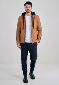 Uomo in piedi con giacca marrone con zip e cappuccio nero, maglietta bianca, pantaloni neri e stivali neri, su sfondo grigio uniforme.