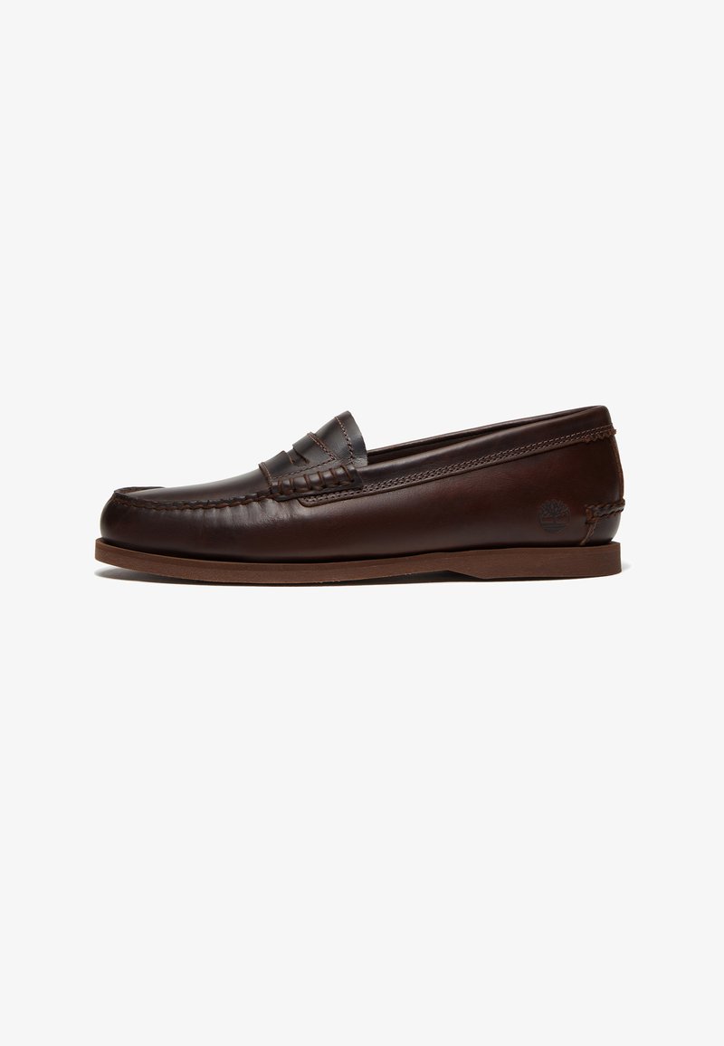 Καφέ δερμάτινο παπούτσι τύπου penny loafer με ραφή moc toe, χαμηλό επίπεδο τακούνι και ανάγλυφο λογότυπο στο πίσω μέρος του παπουτσιού.