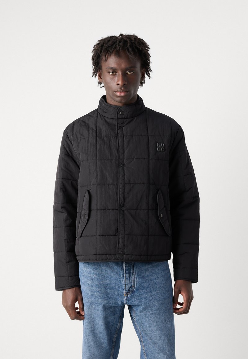 Veste rembourrée noire avec un col montant, des boutons-pression, un texture matelassée et des poches latérales. Présente un logo subtil sur la poitrine.
