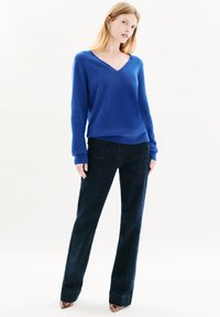 Maglione blu a V in morbido materiale, abbinato a pantaloni in denim scuri con gamba dritta e tasche posteriori sottili. Maniche a sbuffo, aderenti e accorciate.
