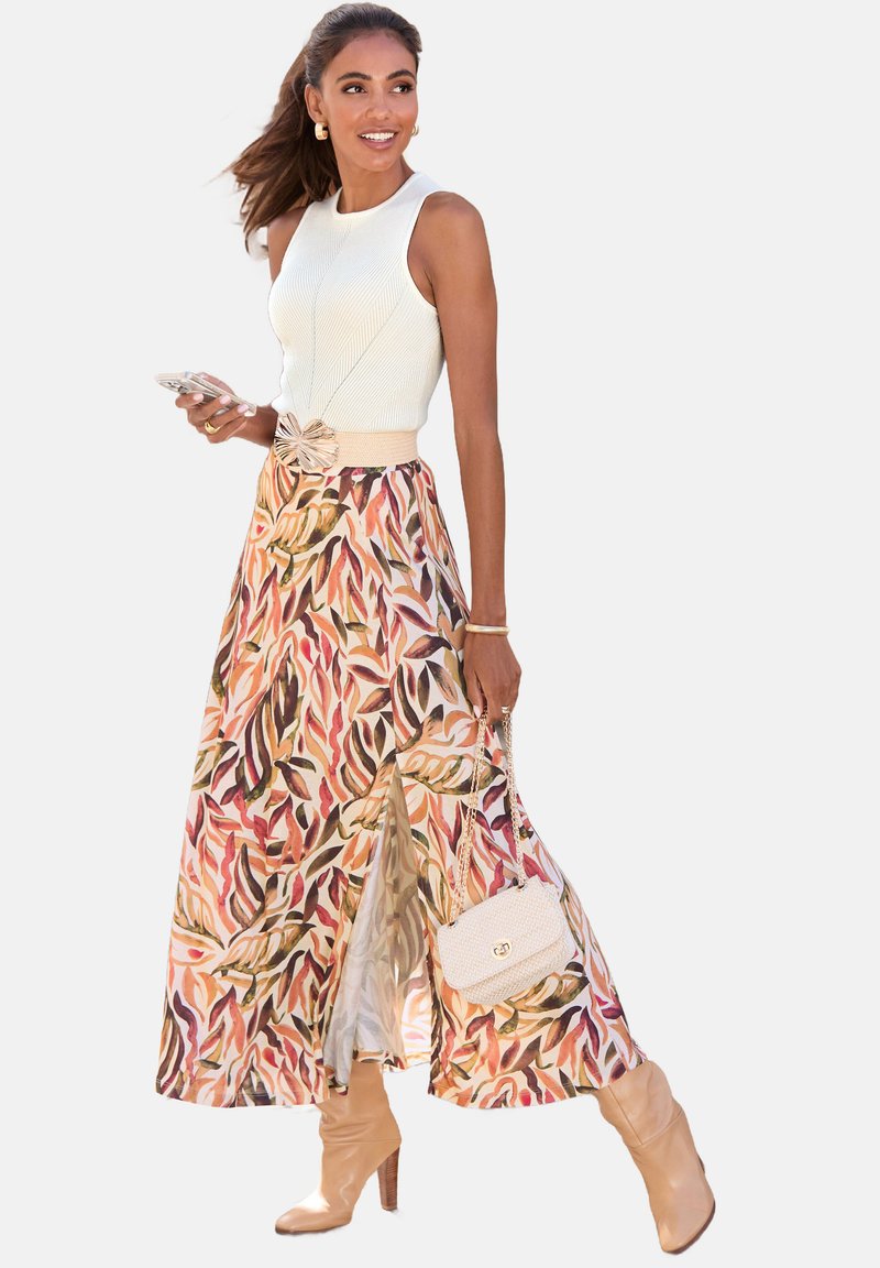 Bjela pletena majica s rebrastom teksturom, maxi suknja s cvjetnim uzorkom u toplim bojama, prorezi i bež čizme na petu; nosi malu bež torbicu.