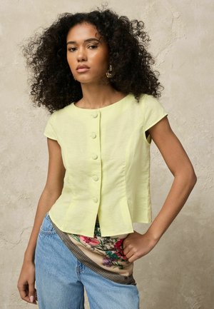 Göndör hajú nő világos sárga gombos felsőt és kék farmert visel, amelybe virágos sál van betűrve, és egy texturált falnak támaszkodik.