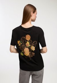 Czarna koszulka z kwiatowym wzorem z tyłu, z przedstawieniem słoneczników i innych kwiatów w różnych kolorach i kształtach. Krótkie rękawy, luźny krój.