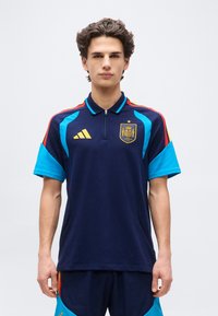 Jaunuolis dėvi tamsiai mėlynį Ispanijos futbolo marškinėlį su geltonu Adidas logotipu, komandos herbu ir šviesiai mėlynomis bei raudonomis peties detalėmis.