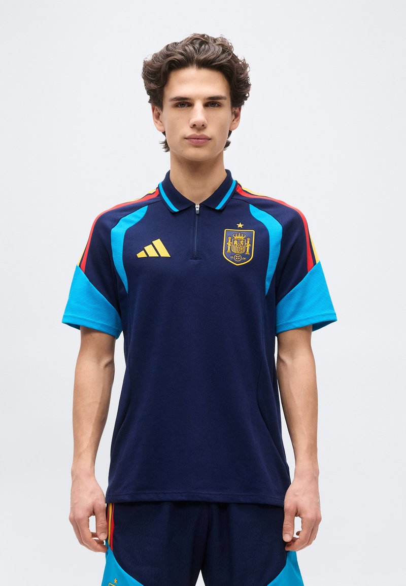 Jaunuolis dėvi tamsiai mėlynį Ispanijos futbolo marškinėlį su geltonu Adidas logotipu, komandos herbu ir šviesiai mėlynomis bei raudonomis peties detalėmis.