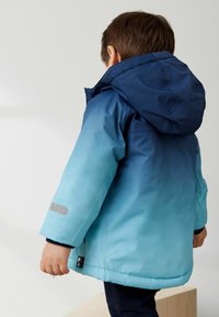 Veste pour enfants passant du bleu marine à la capuche au bleu clair. Fabriquée en tissu brillant avec un accent gris sur la manche.