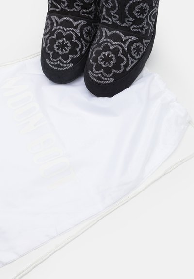 Black suede boots with intricate silver floral embroidery on top, placed on a white drawstring bag with "MOON BOOT" logo.