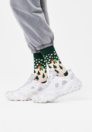 Grønne strømper med høytidsmønstre av trær og røde detaljer, iført med hvite joggesko og grå sweatpants. Detalj av en uformell antrekk.