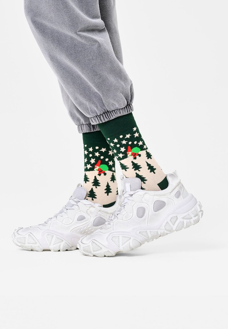 Groene sokken met feestelijke patronen van bomen en rode accenten, gedragen met witte sportschoenen en grijze sweatpants. Casual outfit detail.