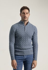 Suéter de punto trenzado en suave lana azul, con cuello alto y tres botones, y puños y dobladillo de canalé. Combinado con jeans azules.