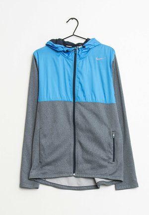 Chaqueta para hombre con parte superior azul y parte inferior gris. Hecha de material ligero, cuenta con un cierre frontal, capucha y bolsillos laterales con cremallera.