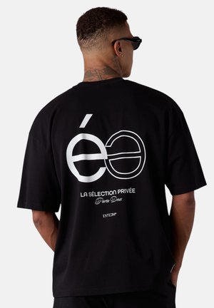 Homme avec un cou tatoué portant des lunettes de soleil noires et un t-shirt noir avec le texte et le logo blancs "éë La Sélection Privée Partie Deux" au dos.