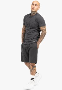 Graues T-Shirt mit kurzen Ärmeln und strukturiertem Logo, kombiniert mit passender Shorts und weißen Sneakers. Hat Tattoos an den Armen.