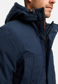 Mørkeblå isolert jakke med teksturert hette, glidelås og trykknapper; har justerbar hette og kontrasterende oransje detaljer.