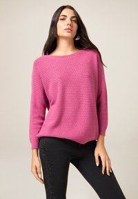 Różowy sweter z dzianiny o luźnym kroju, z okrągłym dekoltem i rękawami do połowy przedramienia. Noszony na czarnych obcisłych dżinsach z ozdobnymi koralikami.